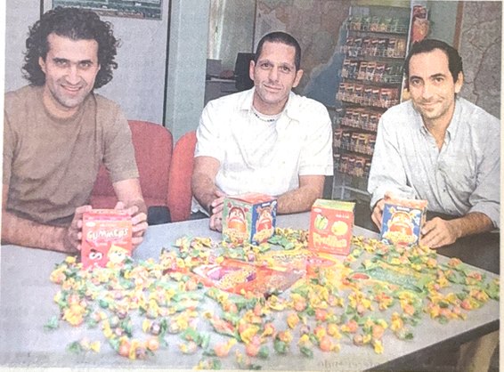 Três homens sentados à mesa com caixas e chicles coloridos espalhados